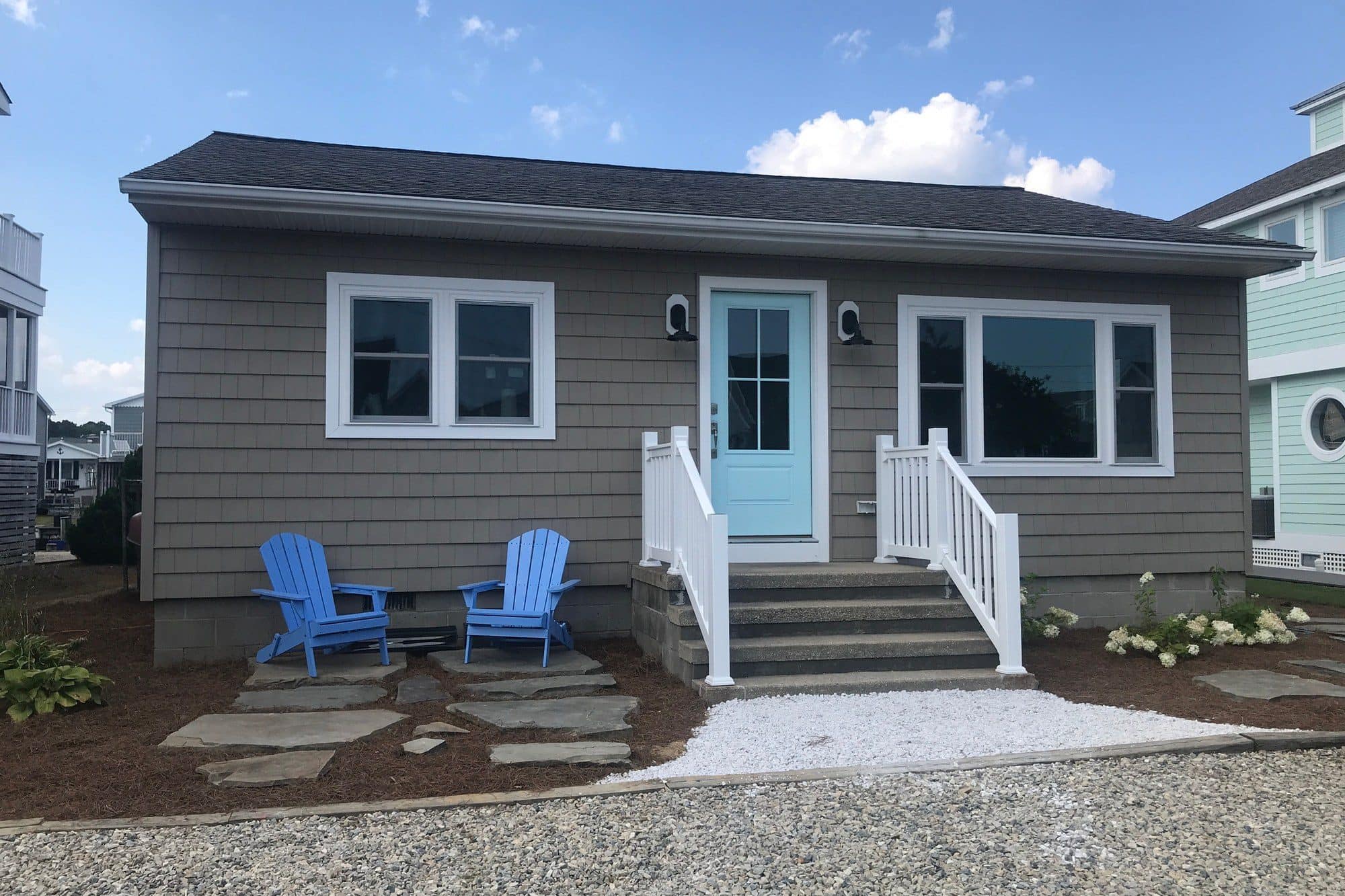 After: the same cottage in gray shingle siding with white trim, an aqua door, and blue Adirondack chairs