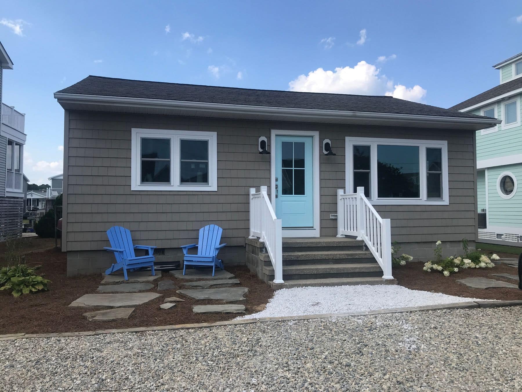 The finished cottage in gray shingle siding with an aqua front door and blue Adirondack chairs